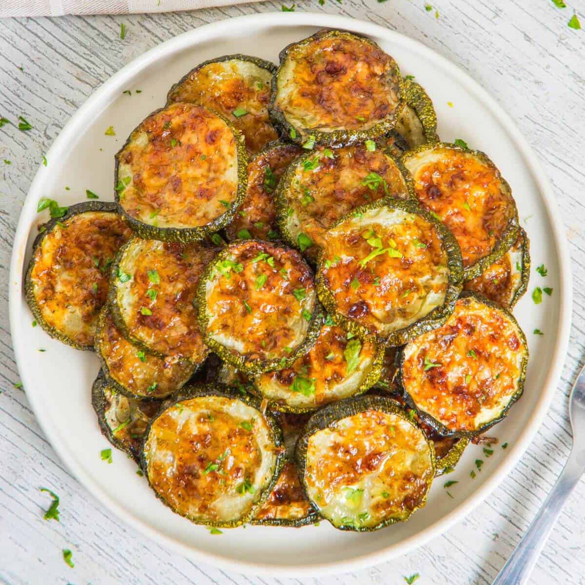 A plate of roasted zucchini slices, lightly browned and garnished with chopped herbs, is set on a white wooden table. A fork is partially visible on the right.
