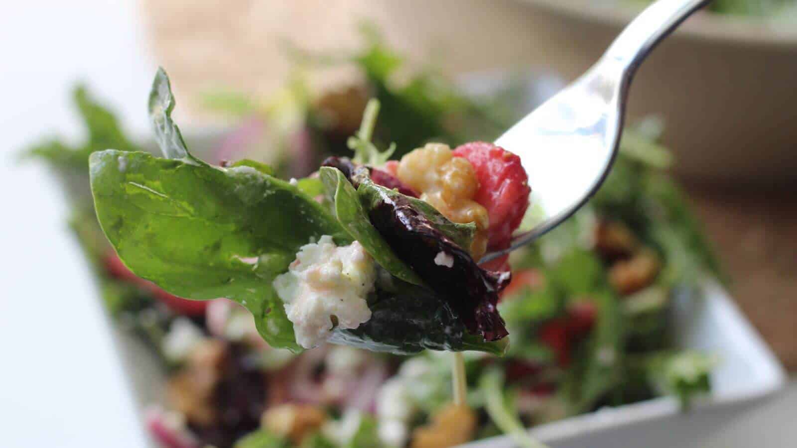 A fork holds a fresh salad with mixed greens, cherry tomato, walnuts, and crumbled cheese over a bowl of salad in the background.