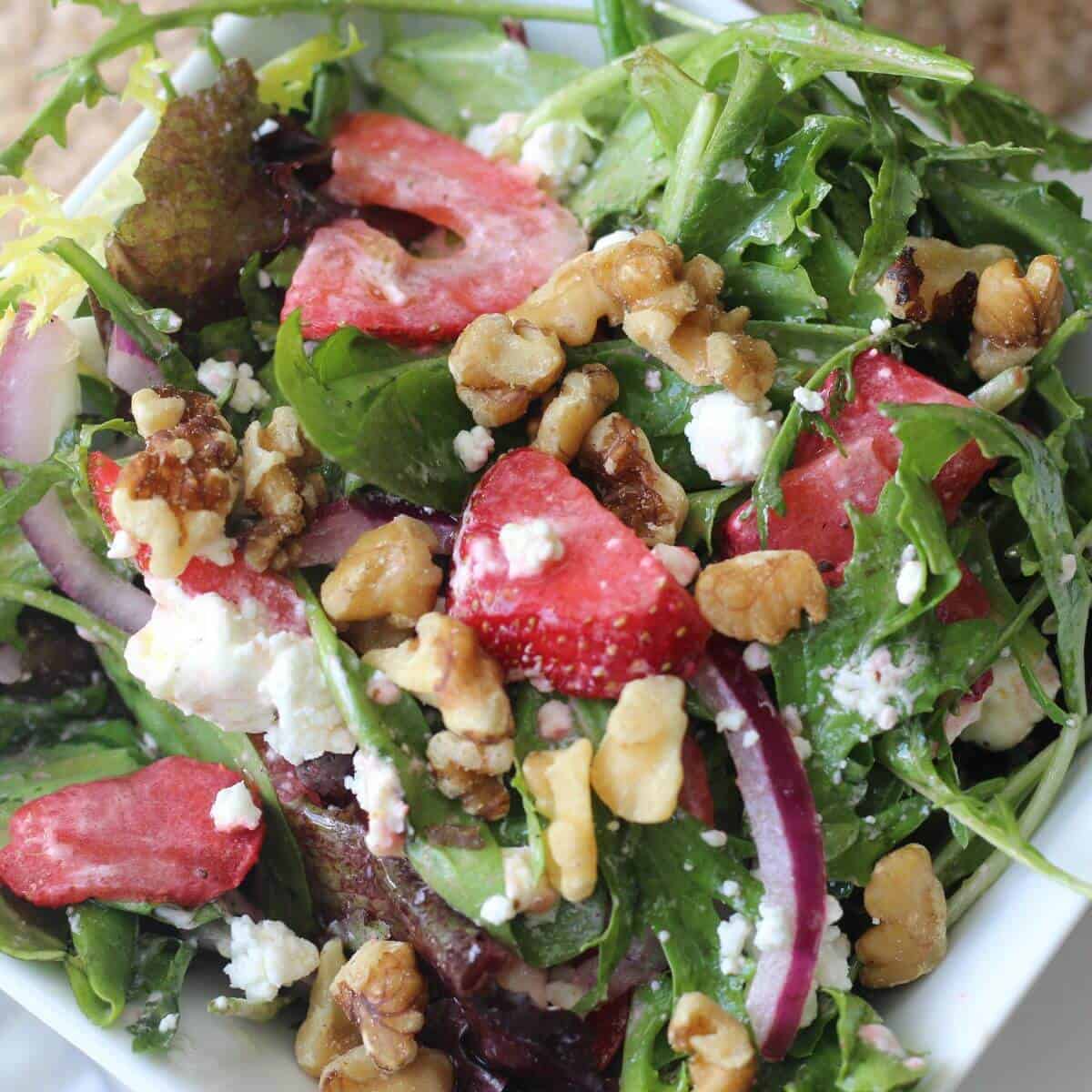 A fresh salad with mixed greens, sliced strawberries, walnuts, red onion, and crumbled cheese in a white bowl.