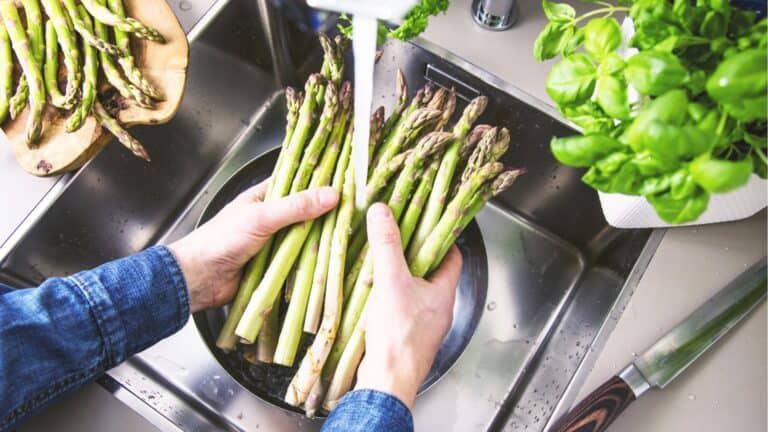 Hands rinsing a bunch of asparagus under running water in a kitchen sink, with a basil plant and knife nearby, showcasing cooking habits to quit for better kitchen practices.