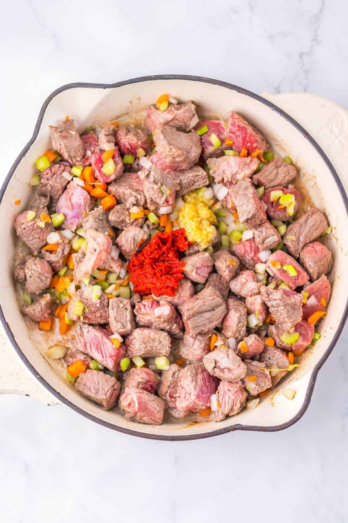 A skillet with diced beef, chopped vegetables, a dollop of tomato paste, and minced garlic on a marble surface.