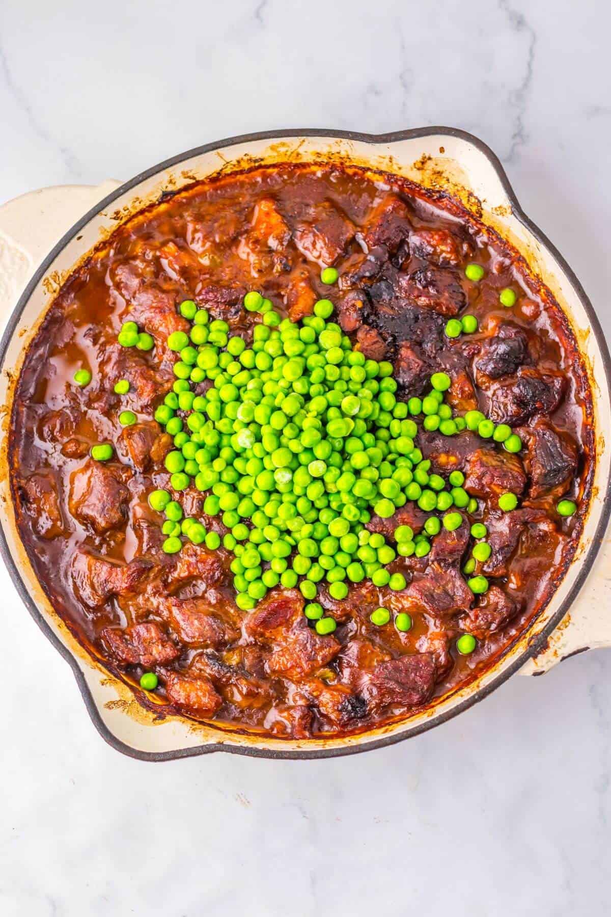 A pot of stew with chunks of meat in rich sauce, topped with a pile of green peas, on a marble countertop.