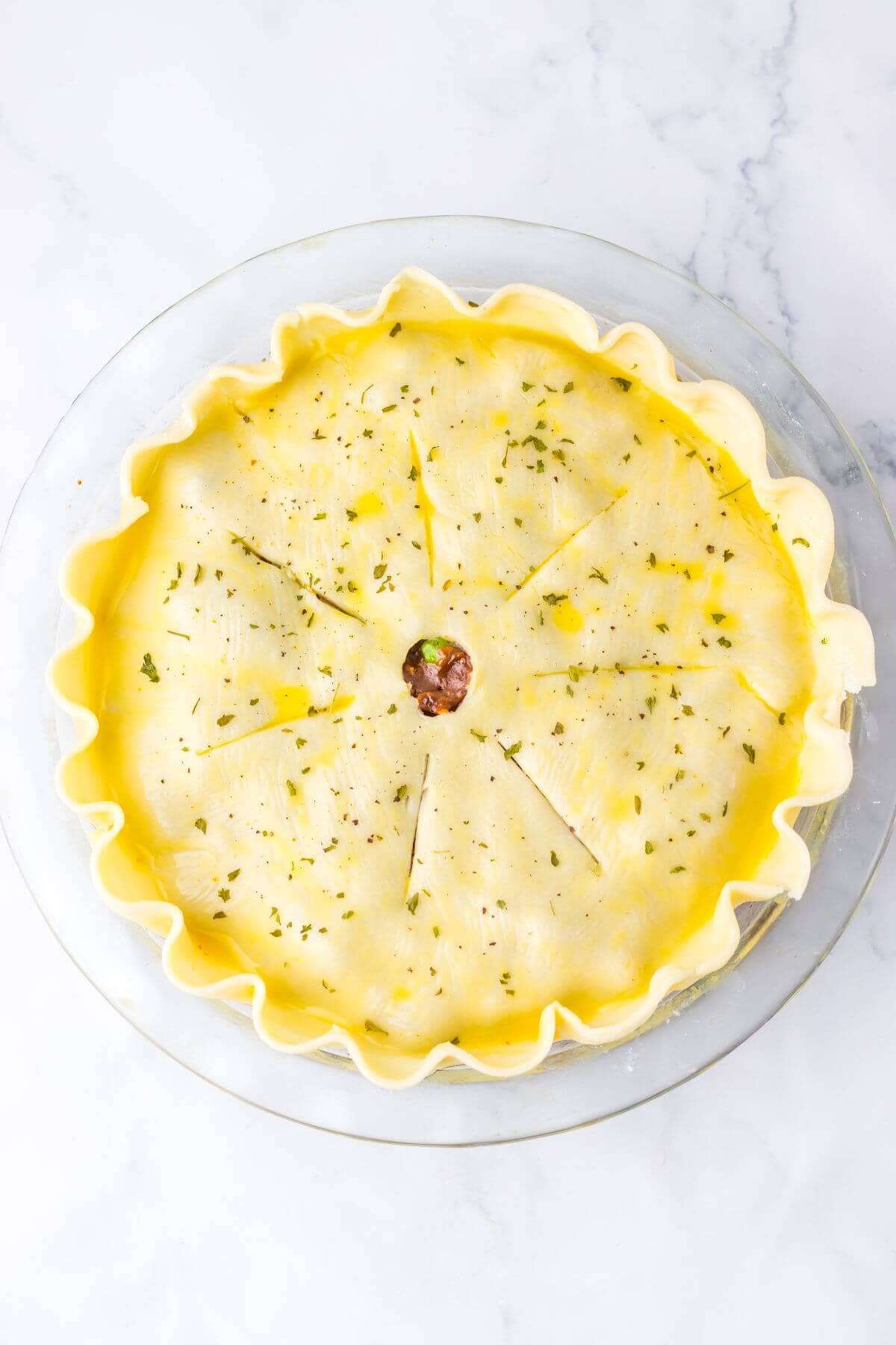 A pie with a decorative crust, featuring slits and a small central vent, is ready for baking on a round glass dish.