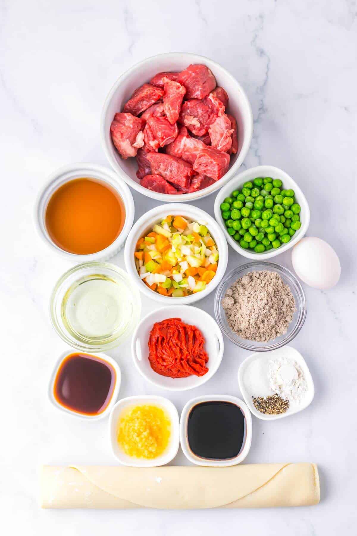 Various cooking ingredients are arranged on a white surface, including diced meat, peas, diced vegetables, sauces, flour, egg, tomato paste, stock, and a pastry sheet.