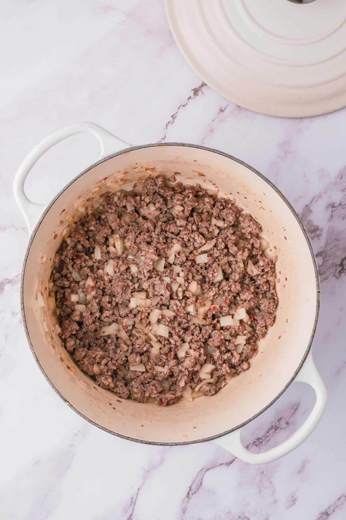 A white pot containing cooked ground beef and chopped onions on a marble surface.