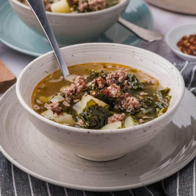A bowl of soup with chunks of meat, leafy greens, and potato cubes, served with a spoon on a stacked bowl and plate set.