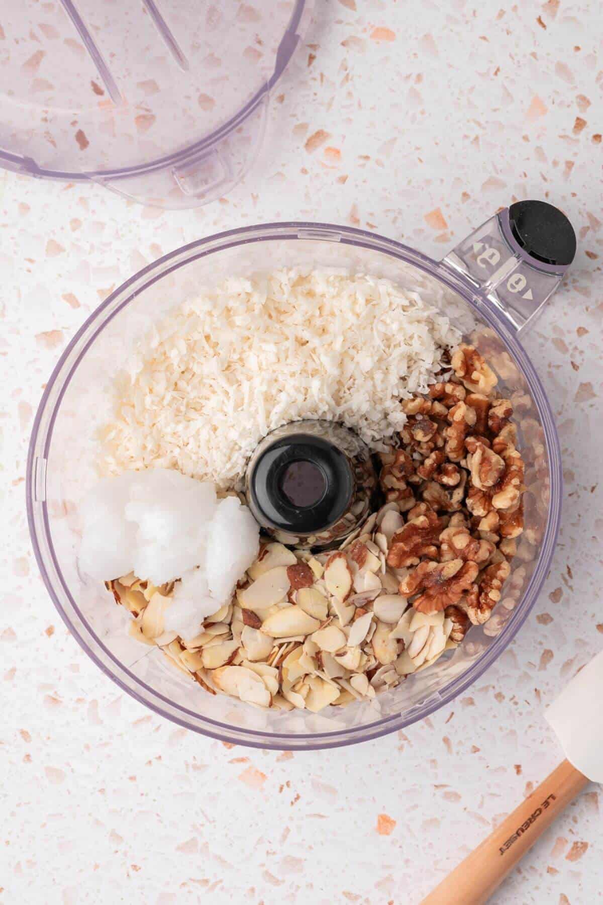 A food processor bowl containing coconut flakes, walnuts, sliced almonds, and coconut oil on a speckled countertop.
