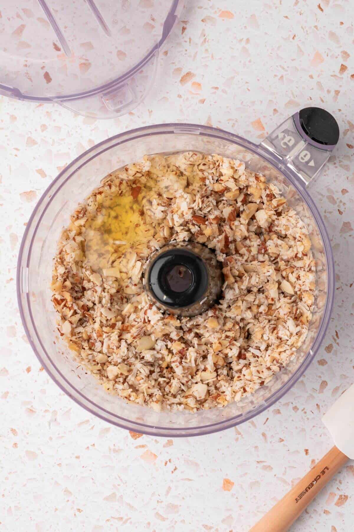 Chopped nuts and oil in a food processor bowl, viewed from above, on a speckled countertop.
