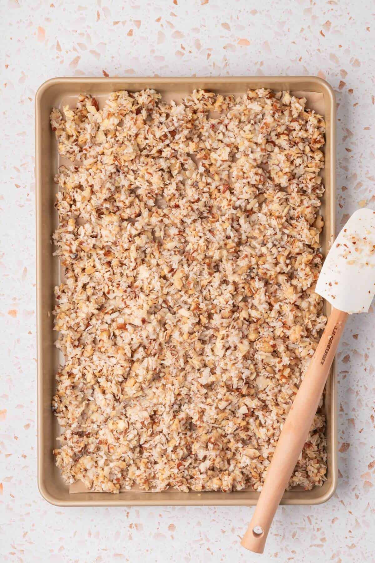 A baking sheet with a layer of granola, featuring oats and nuts, and a spatula resting on the side.