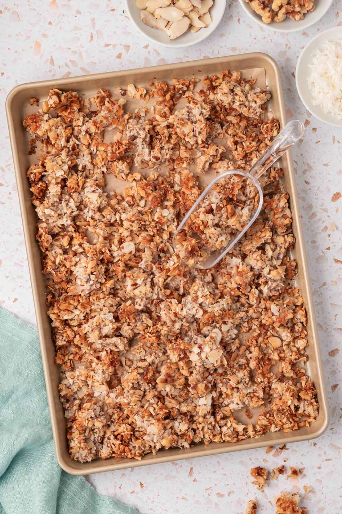 A baking tray filled with toasted granola, accompanied by a clear serving scoop. Two small bowls with nuts and a green cloth are on the side.