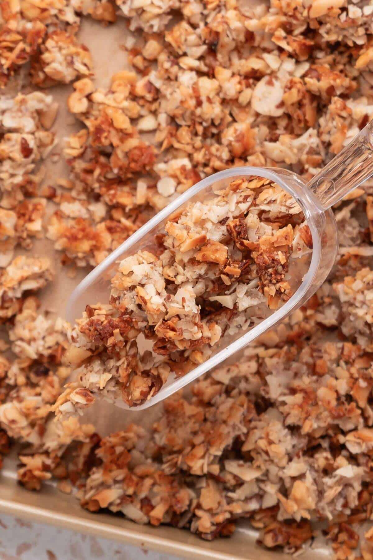 A scoop of granola with oats, nuts, and seeds resting on a pile of the same mixture on a baking sheet.