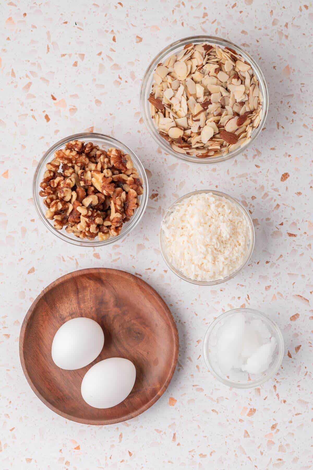 Bowls of sliced almonds, chopped walnuts, shredded coconut, two eggs on a wooden plate, and a small bowl of coconut oil on a speckled surface.