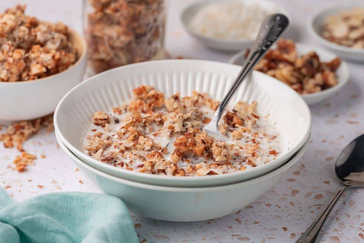 A bowl of cereal with milk and a spoon, surrounded by small bowls of granola on a speckled surface.