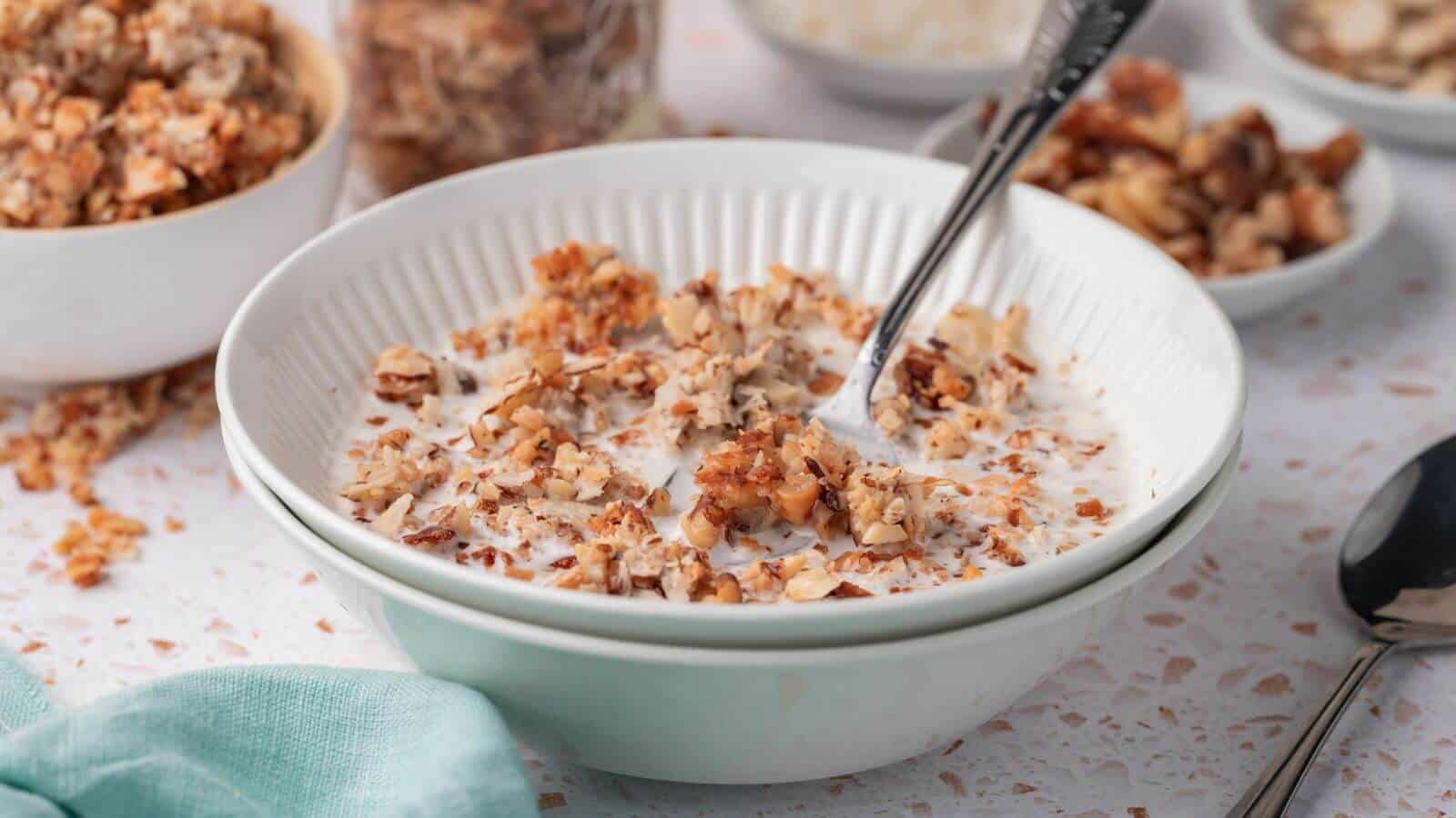 A bowl of granola mixed with milk, with a spoon inside. Other bowls of granola and toppings visible in the background. A teal cloth is partially visible on the side.