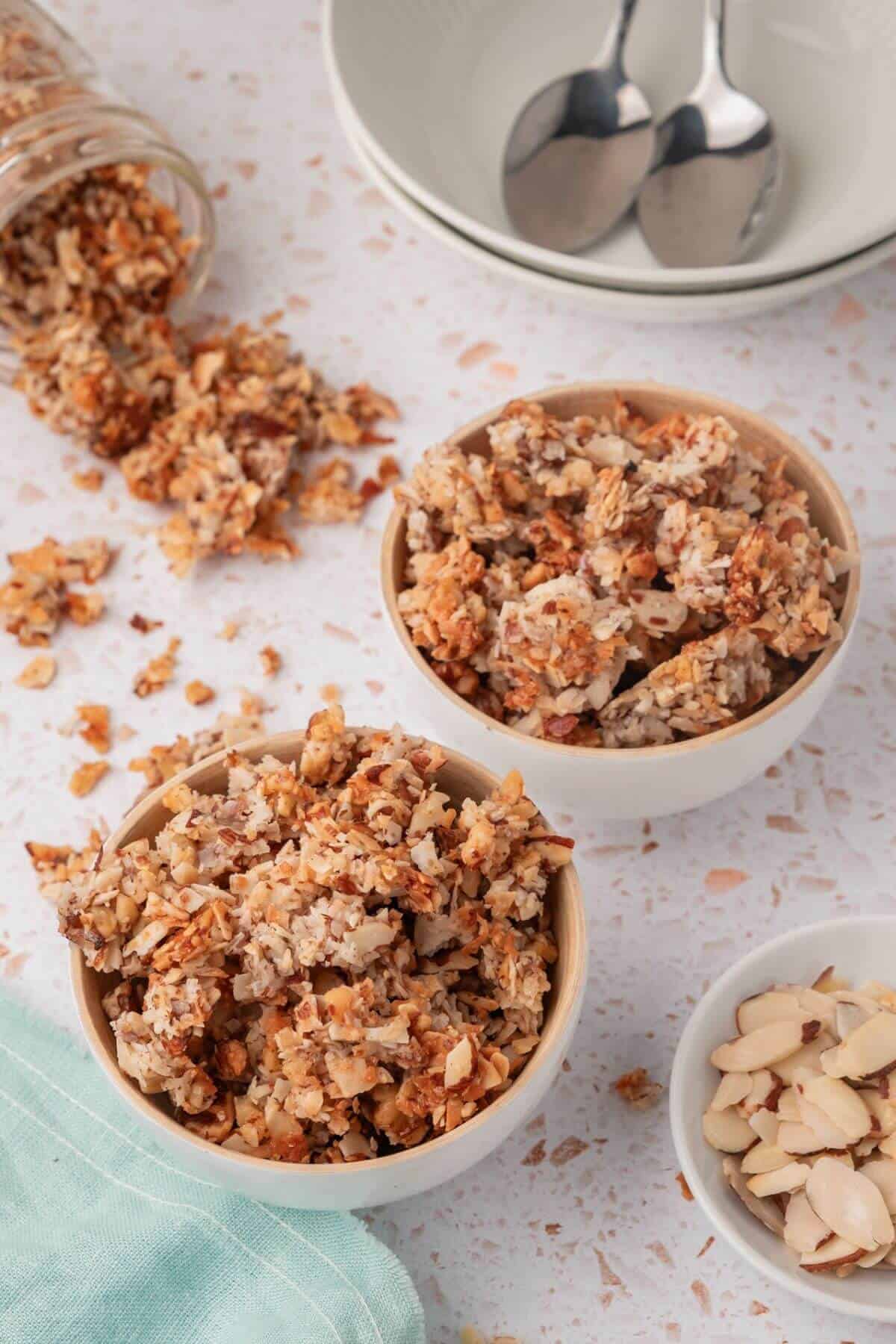 Bowls of granola on a table with scattered granola pieces, a jar, two empty bowls, and spoons nearby.