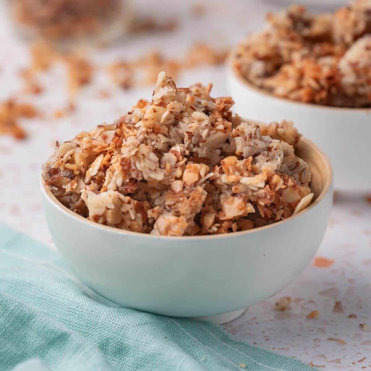 A white bowl filled with homemade nut granola is placed on a light-colored surface, with some granola spilled around the bowl. A folded teal cloth is partially visible beside it.