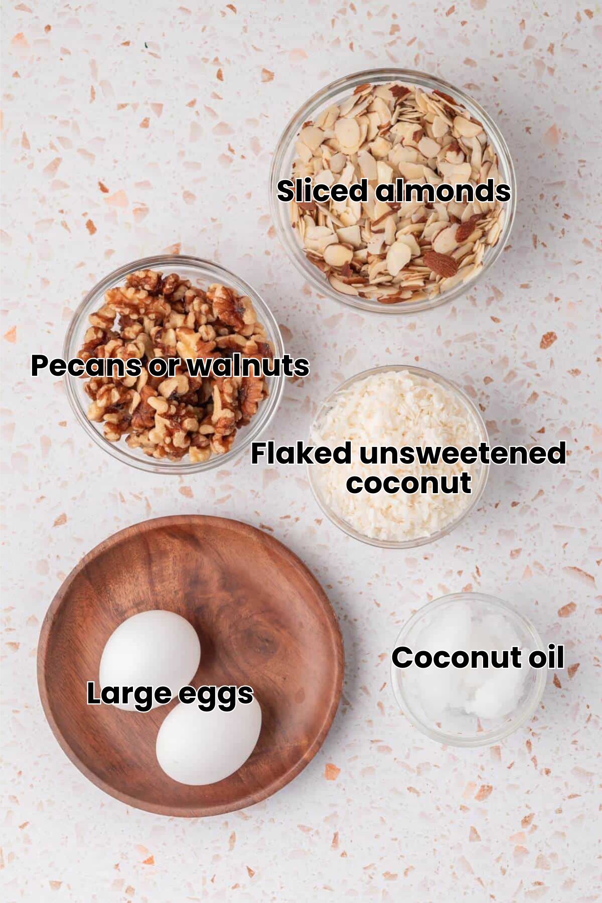 Bowls of sliced almonds, chopped walnuts, shredded coconut, two eggs on a wooden plate, and a small bowl of coconut oil on a speckled surface.