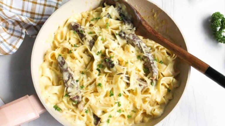 A pan of creamy pasta with sliced beef and parsley, topped with grated cheese. A wooden spoon rests in the pan.