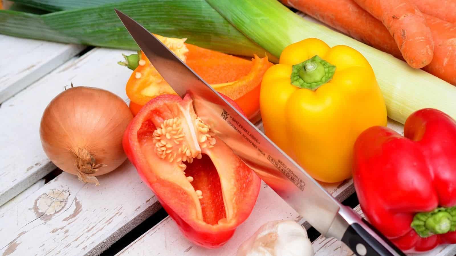 A knife rests on chopped vegetables, including red and yellow bell peppers, an onion, carrots, a leek, and garlic, placed on a white wooden surface.