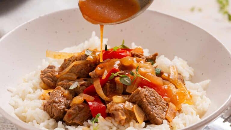 Close-up of a white bowl with rice topped with beef, onions, and red peppers. Sauce is being poured over the dish.