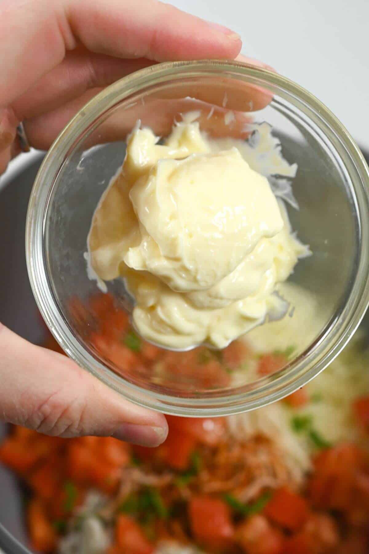 A hand holds a small glass bowl of mayonnaise over chopped tomatoes and herbs in another bowl.