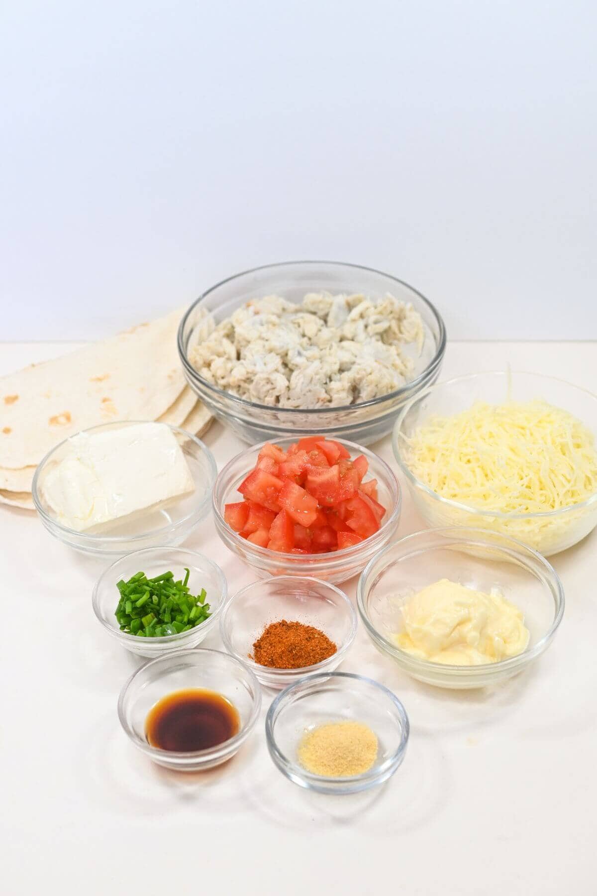 Ingredients arranged on a table include chopped chicken, diced tomatoes, shredded cheese, tortillas, cream cheese, spices, mayonnaise, soy sauce, and chopped green onions in clear bowls.