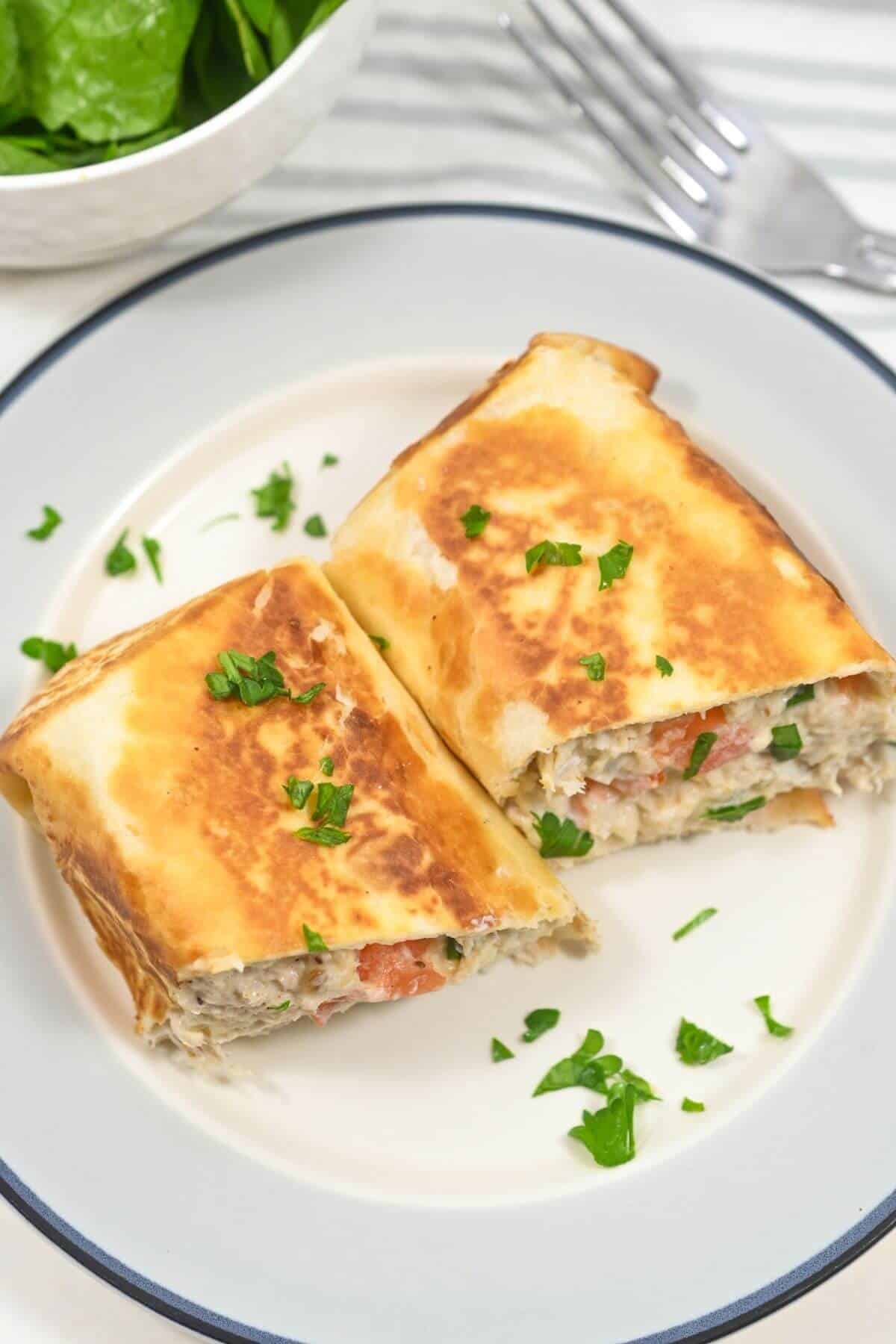 Two golden-brown stuffed wraps on a plate, garnished with chopped parsley. A fork and small salad bowl are in the background.
