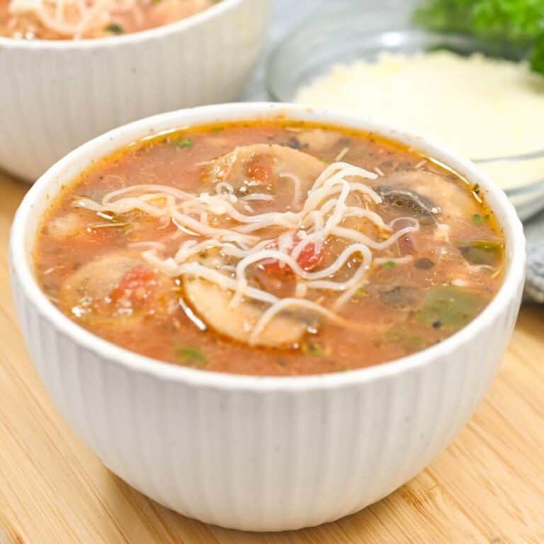 A bowl of vegetable soup with mushrooms, tomatoes, and noodles on a wooden surface. Another bowl and grated cheese are blurred in the background.