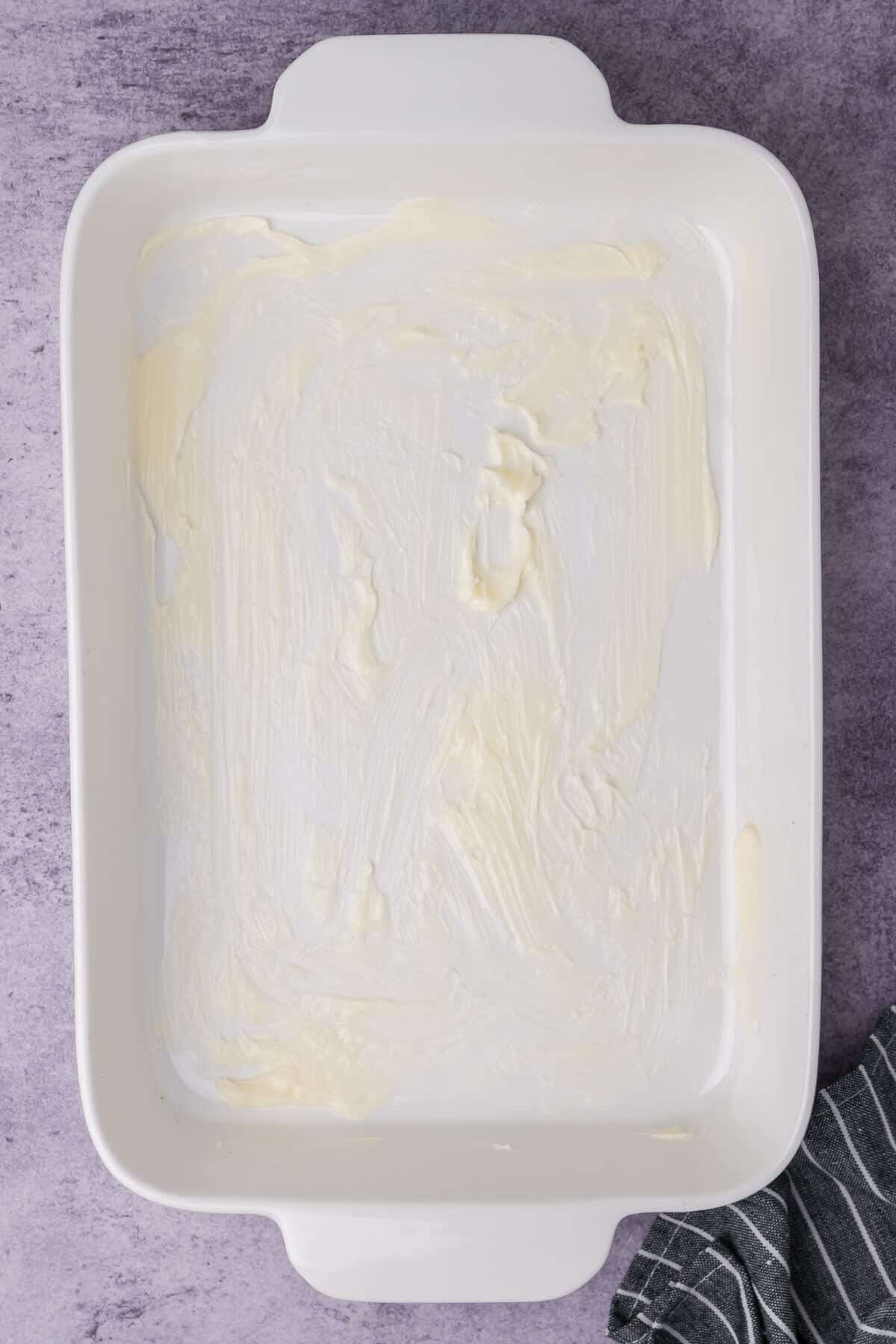 A white baking dish greased with butter on a purple countertop, with a striped cloth partially visible at the bottom right corner.