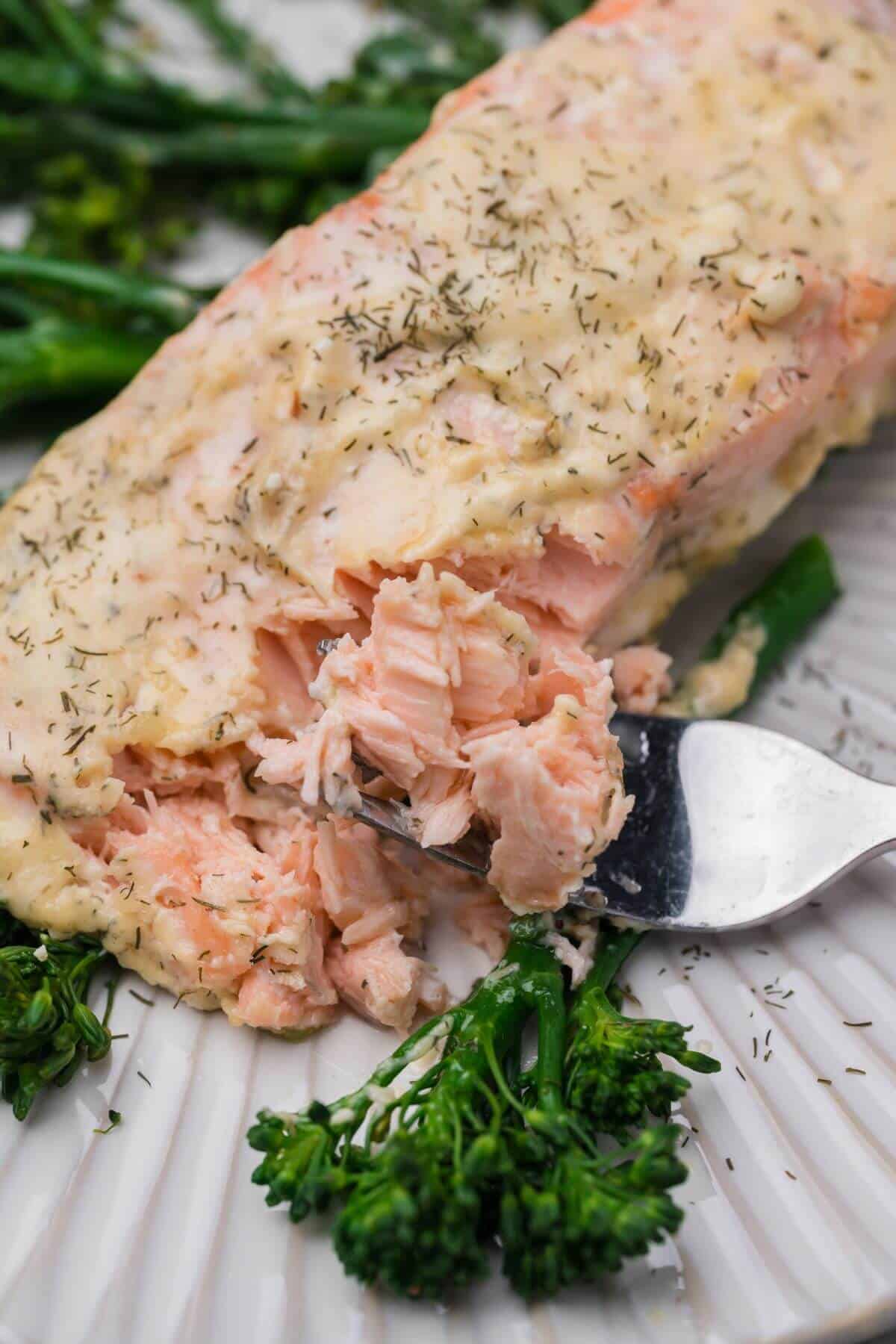 Close-up of a fork cutting into a creamy baked salmon fillet, seasoned with herbs, served on a plate with steamed broccolini.