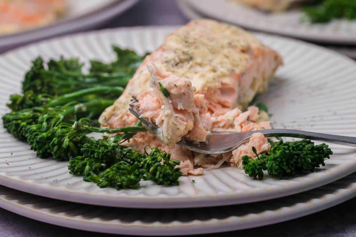A plate with baked salmon topped with herbs and a dollop of cream sauce, accompanied by steamed broccolini. A fork is flaking a piece of the salmon.