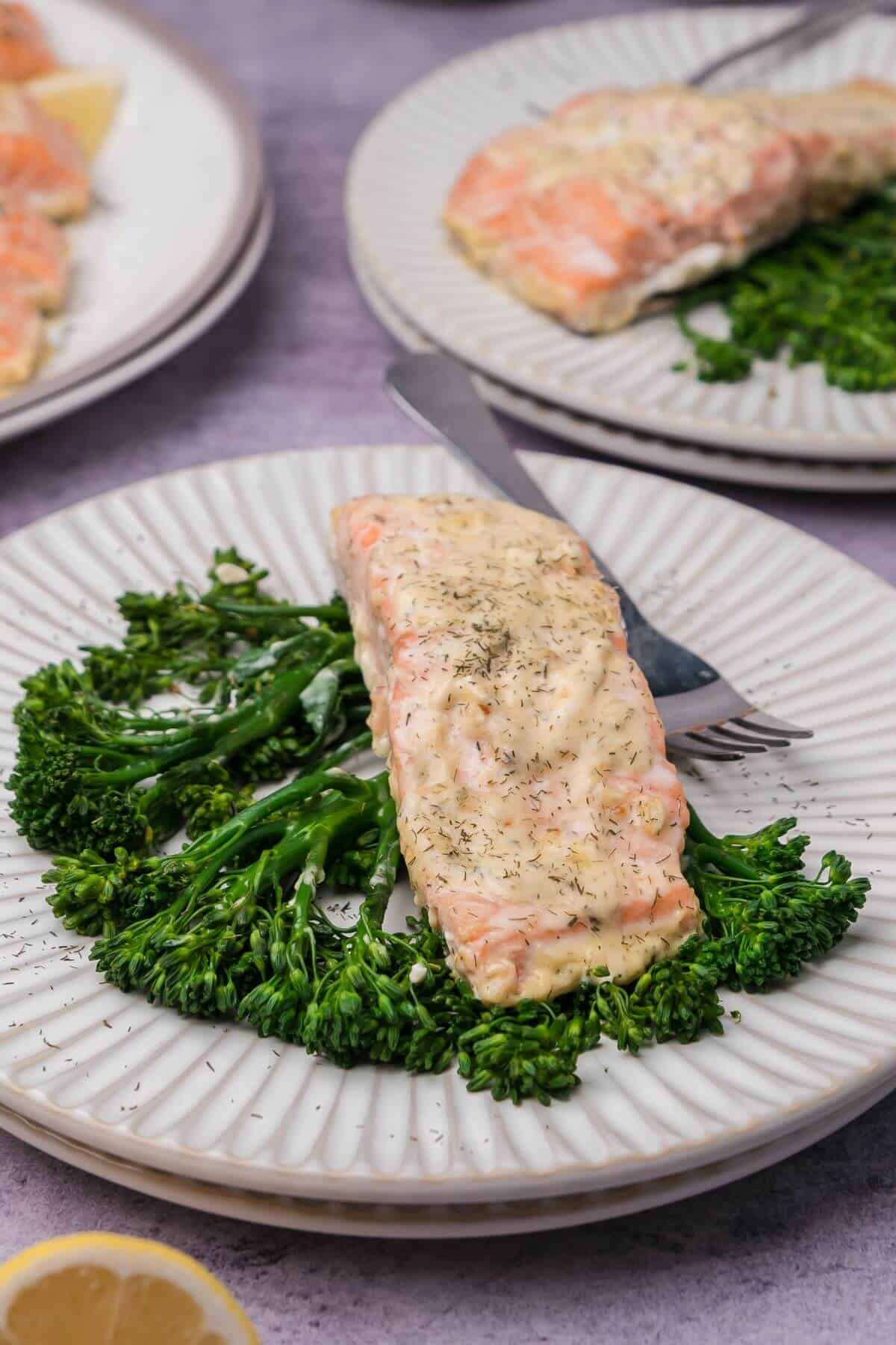 Cooked salmon fillet with creamy dill sauce on a bed of broccolini on a white plate, with a fork and knife beside it.