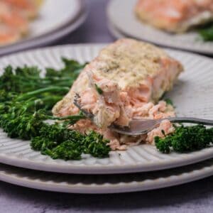Plate of baked salmon with herbs, served with broccolini on a textured white dish. A fork is holding a piece of salmon.