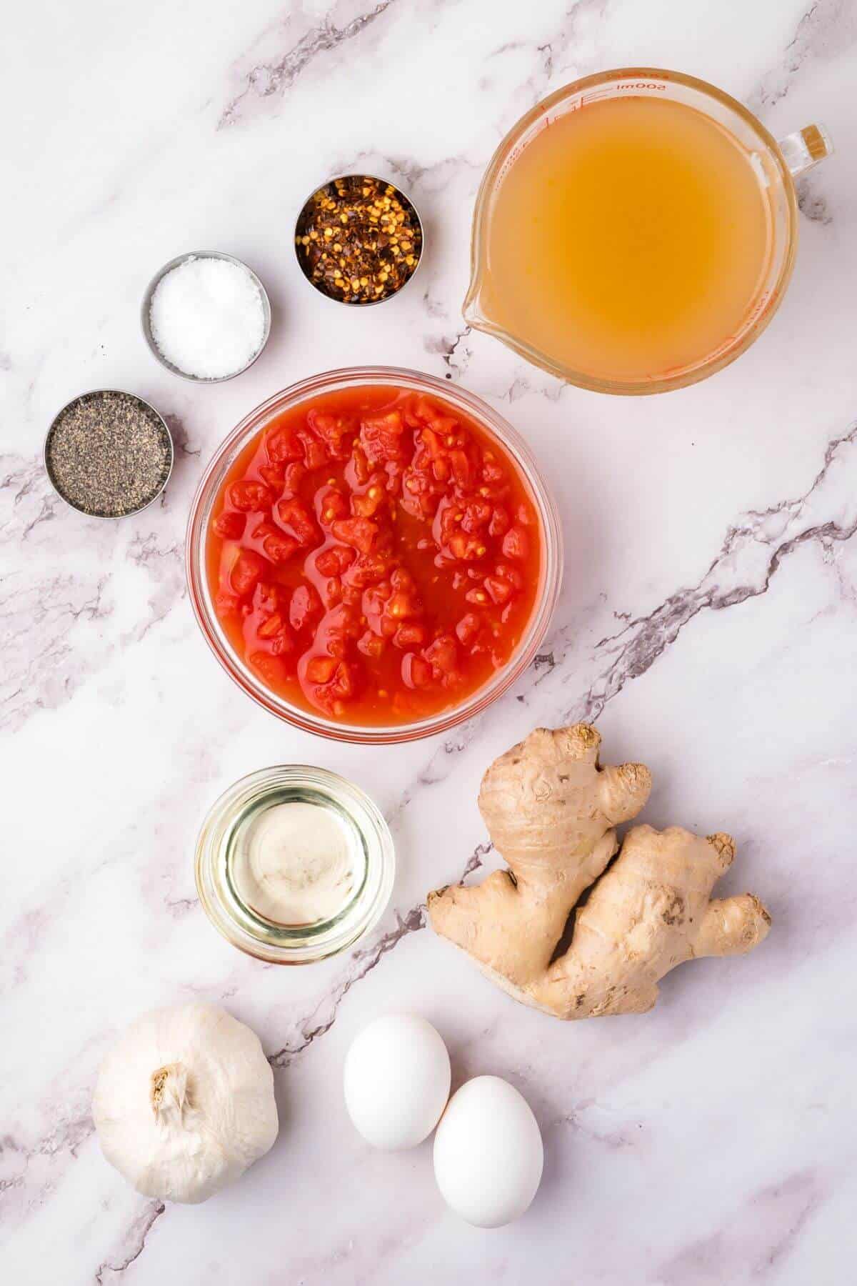 Ingredients on a marble surface: bowl of diced tomatoes, ginger root, two eggs, head of garlic, small bowl of oil, bowls of black pepper, salt, crushed red pepper, and a jug of broth.