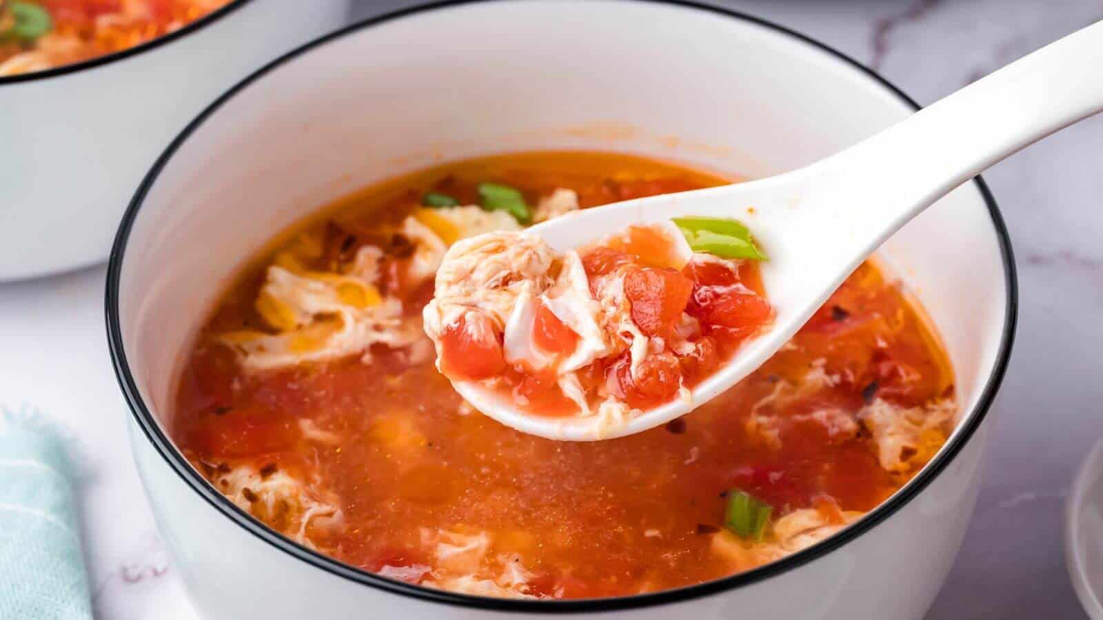 A spoonful of tomato egg drop soup over a bowl, showing diced tomatoes and green onions.