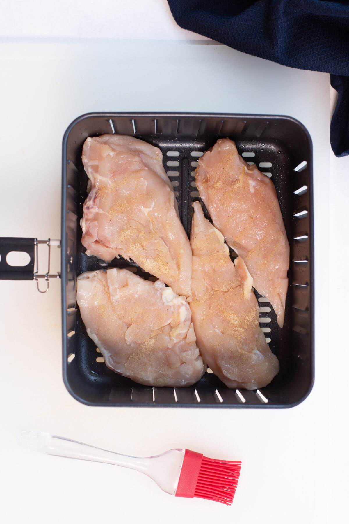 Raw chicken breasts in an air fryer basket with a red basting brush nearby.