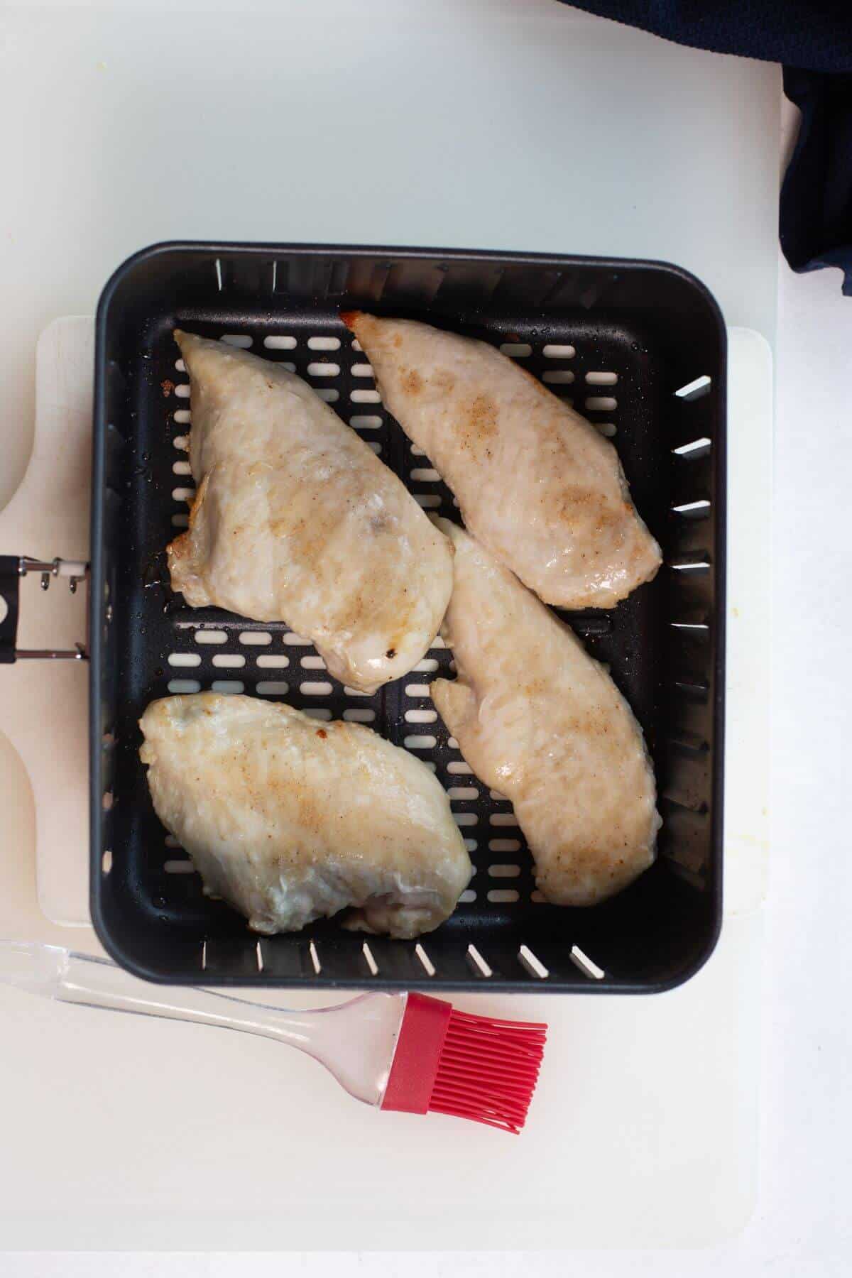 Four cooked chicken breasts in an air fryer basket with a red basting brush on the side.