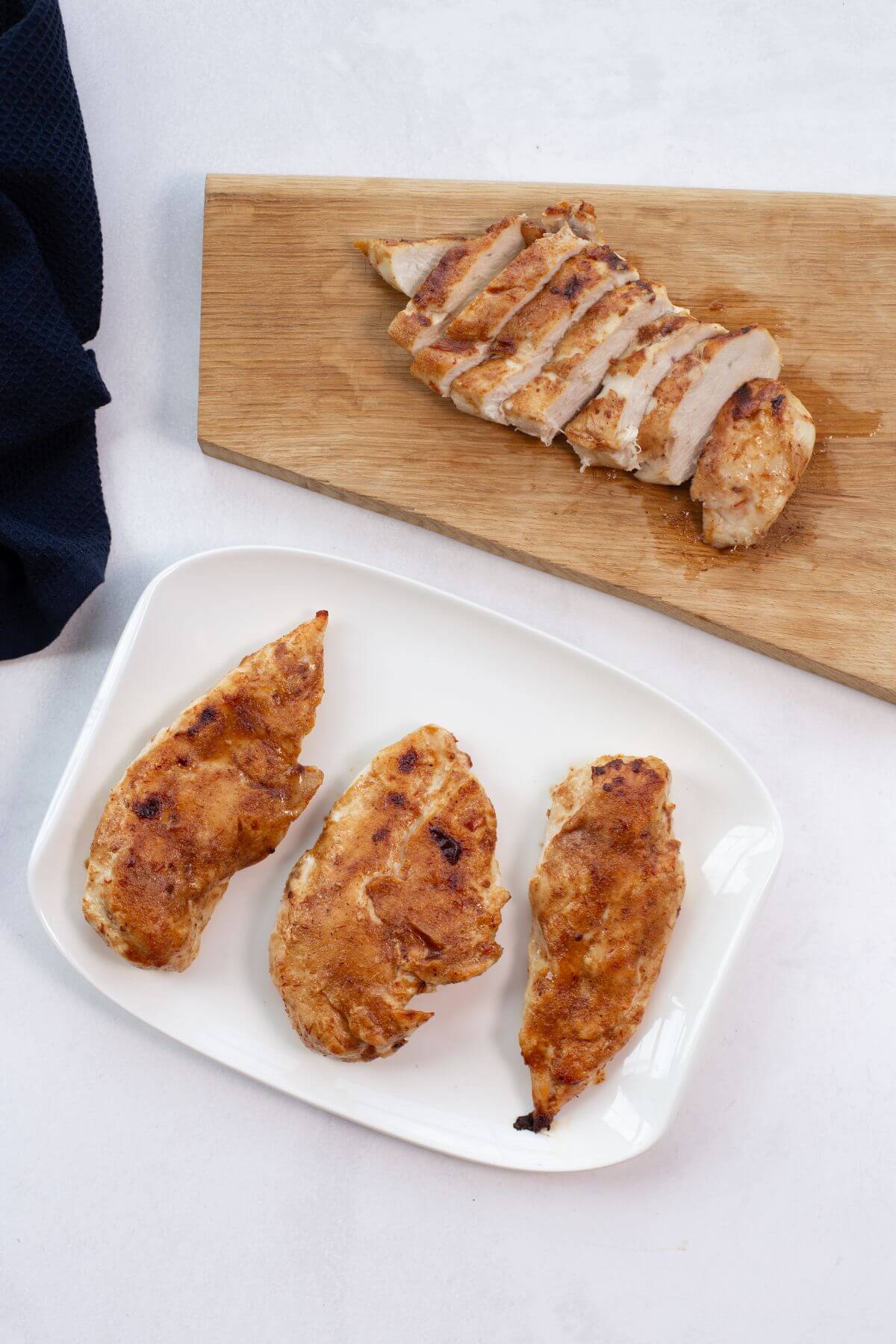 Sliced cooked chicken breast on a wooden board, with three additional pieces on a white plate.
