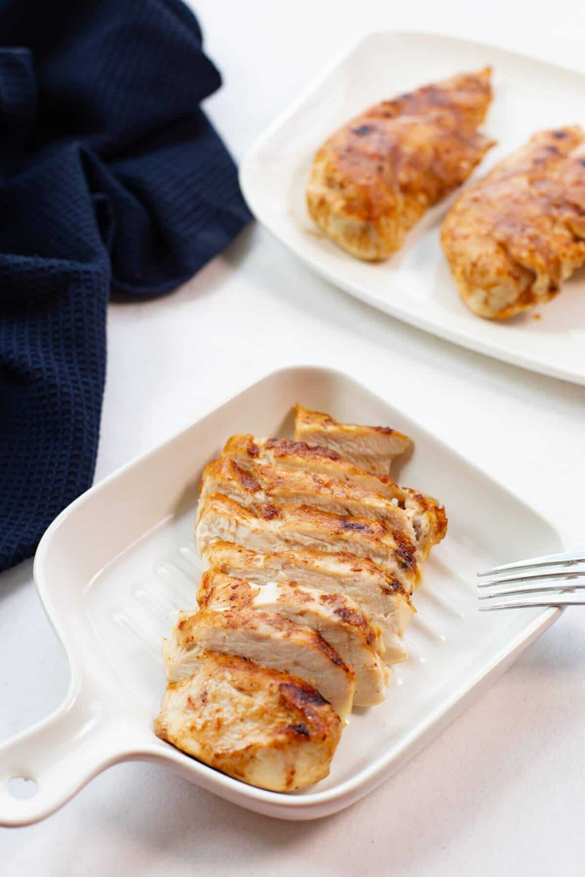 Two plates of grilled chicken breast; one plate has sliced pieces and the other has whole pieces. A fork is resting on the edge of the sliced chicken plate. A dark cloth is beside them.