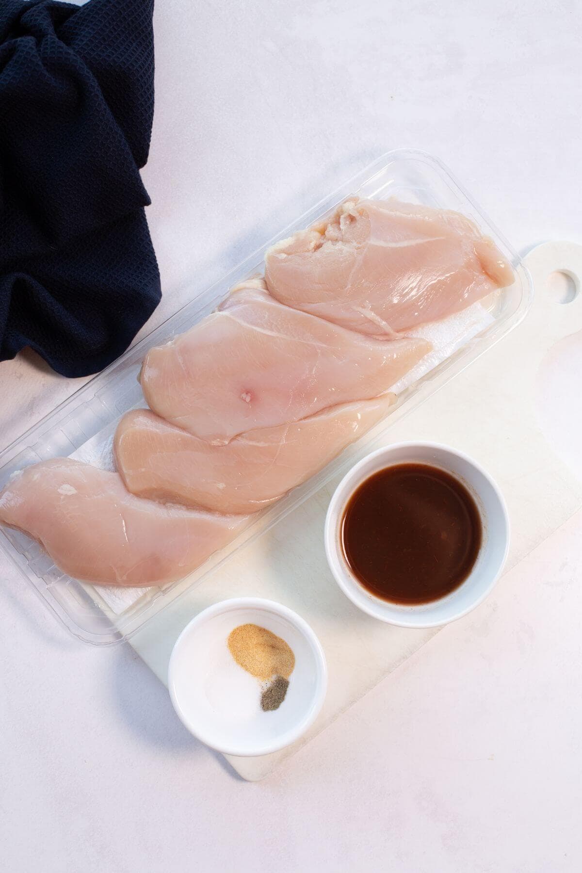 Four raw chicken breasts on a tray beside small bowls of sauce and seasoning on a white cutting board.