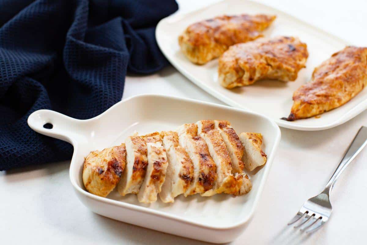 Sliced chicken breast in a ceramic dish with whole chicken pieces on a plate in the background. A fork lies nearby on a white surface.