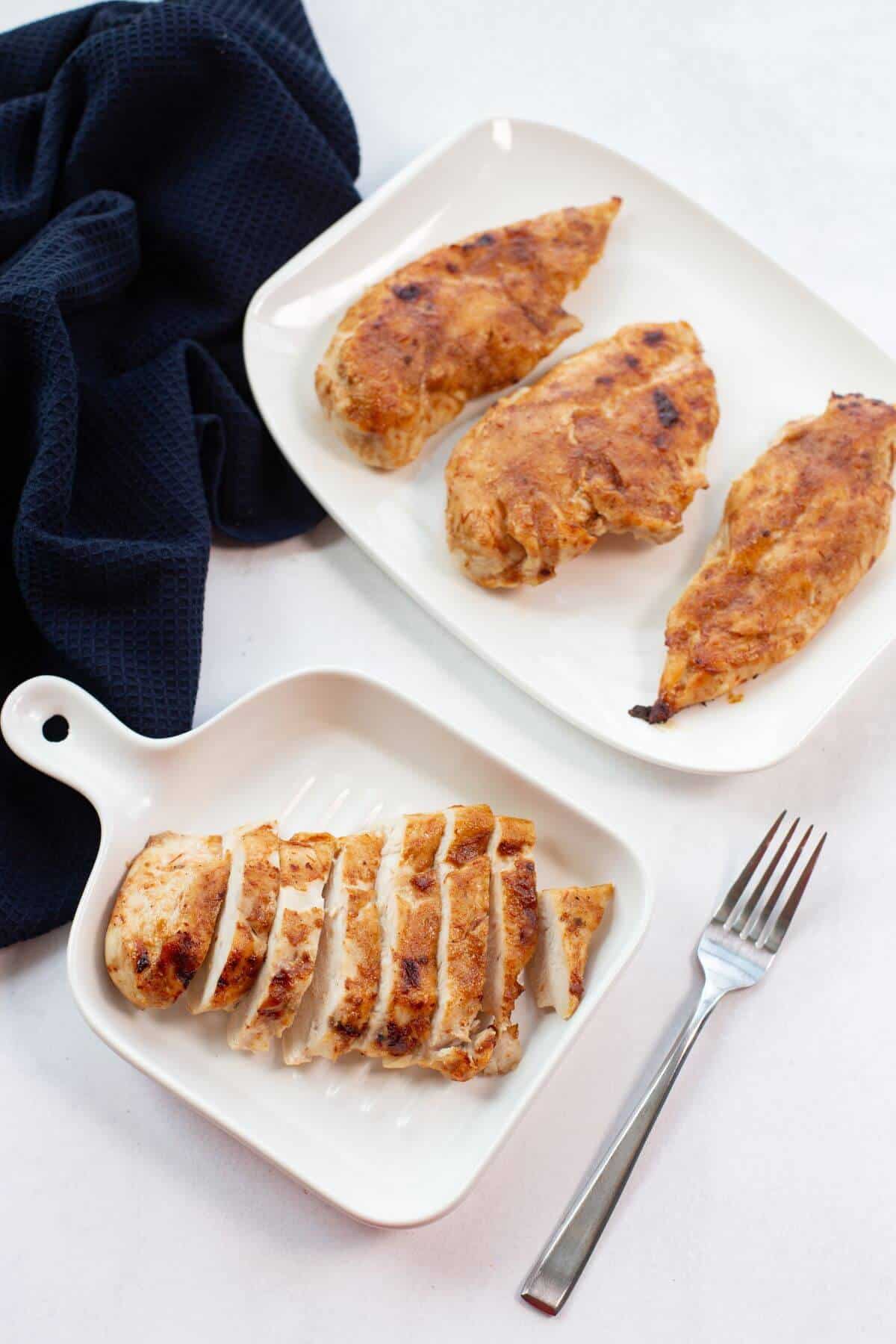 Two white dishes display grilled chicken, one with whole pieces and the other with sliced pieces. A fork lies nearby, and a dark cloth is in the background.