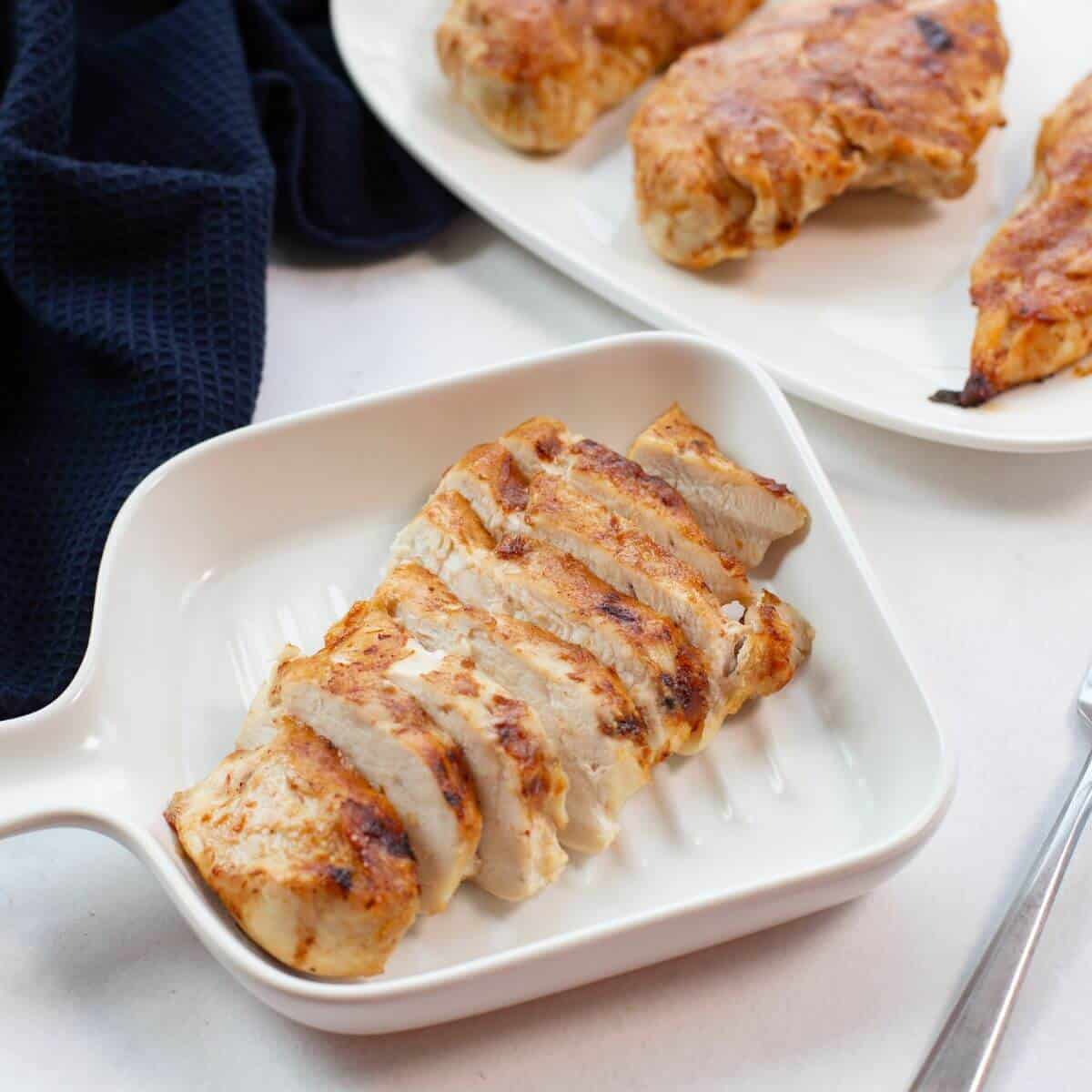 Sliced grilled chicken breast in a white dish, with more chicken pieces on a separate plate in the background. A dark cloth is partially visible.