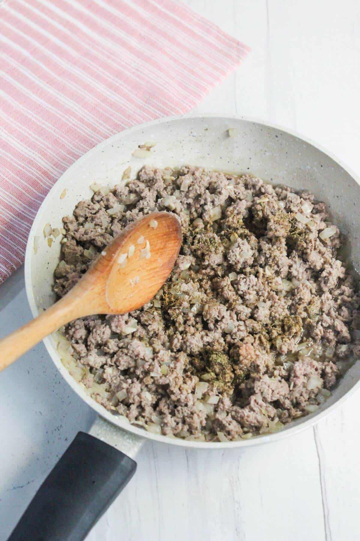 Pan with cooked ground meat and onions, stirred with a wooden spoon, on a light-colored surface next to a red and white striped cloth.