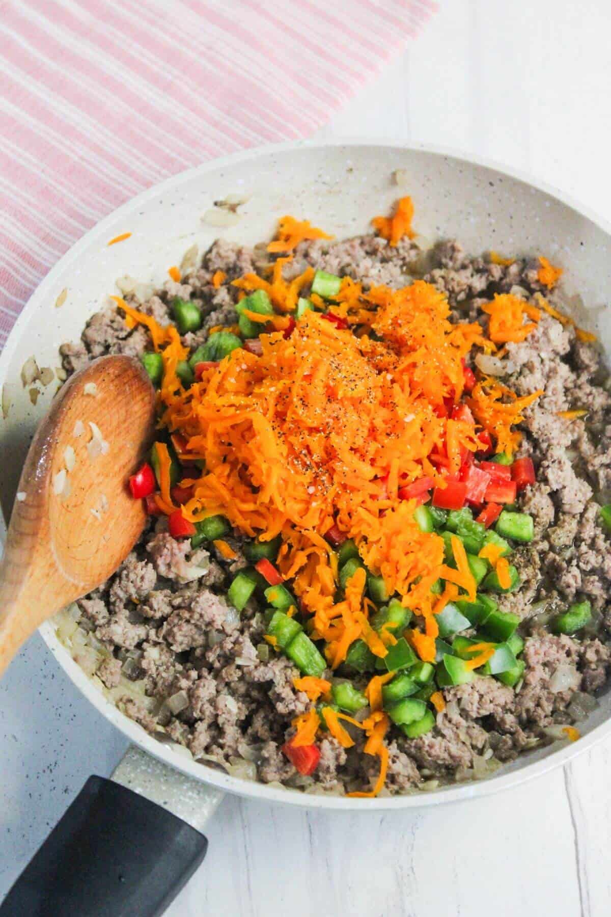 Ground meat, diced green and red bell peppers, grated orange cheese, and a sprinkle of seasoning in a frying pan with a wooden spoon.