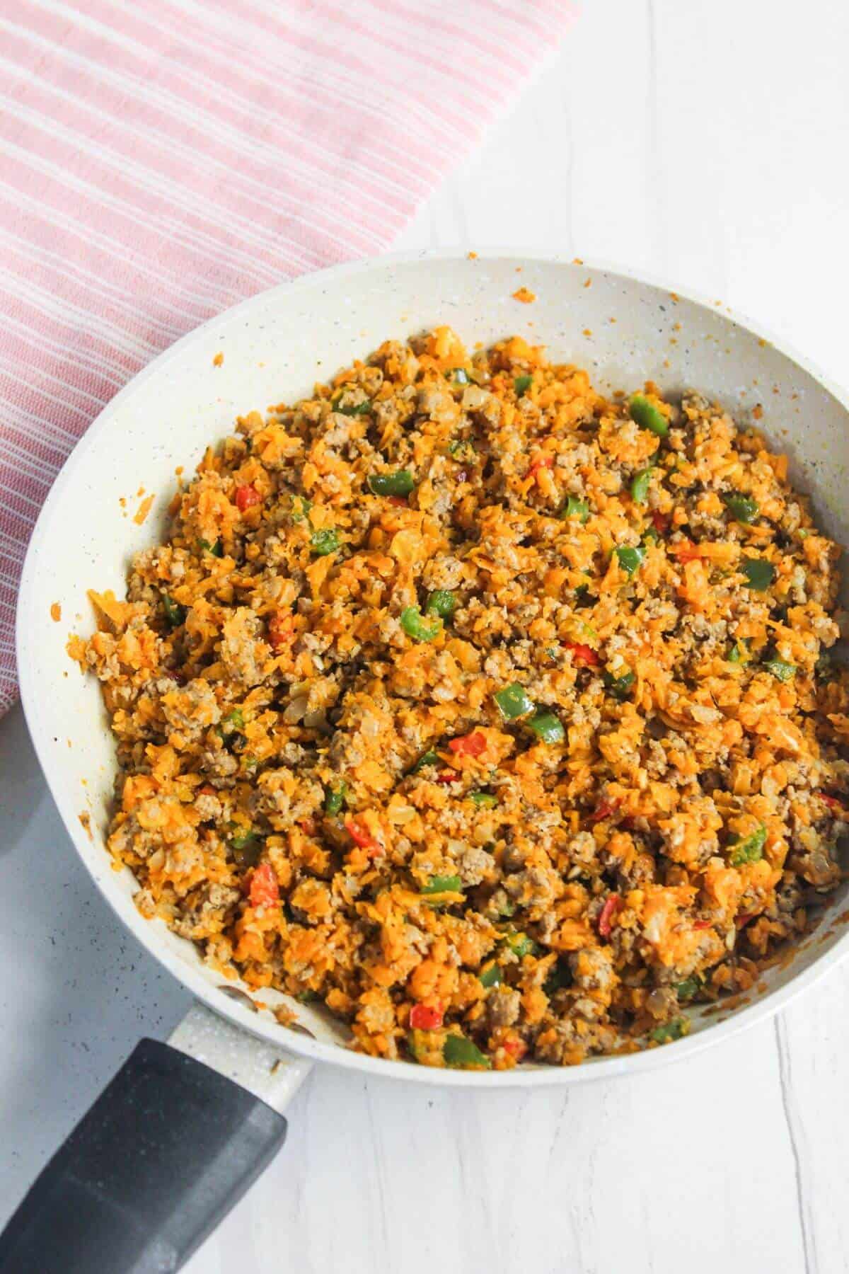 A skillet filled with seasoned cauliflower rice, mixed with green and red bell peppers and spices, placed on a light wooden surface next to a striped cloth.