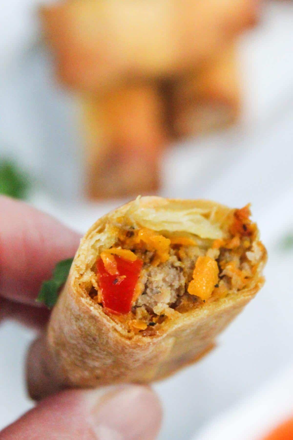 Close-up of a hand holding a spring roll with a visible filling of minced meat, red bell peppers, and vegetables. Other spring rolls are blurred in the background.