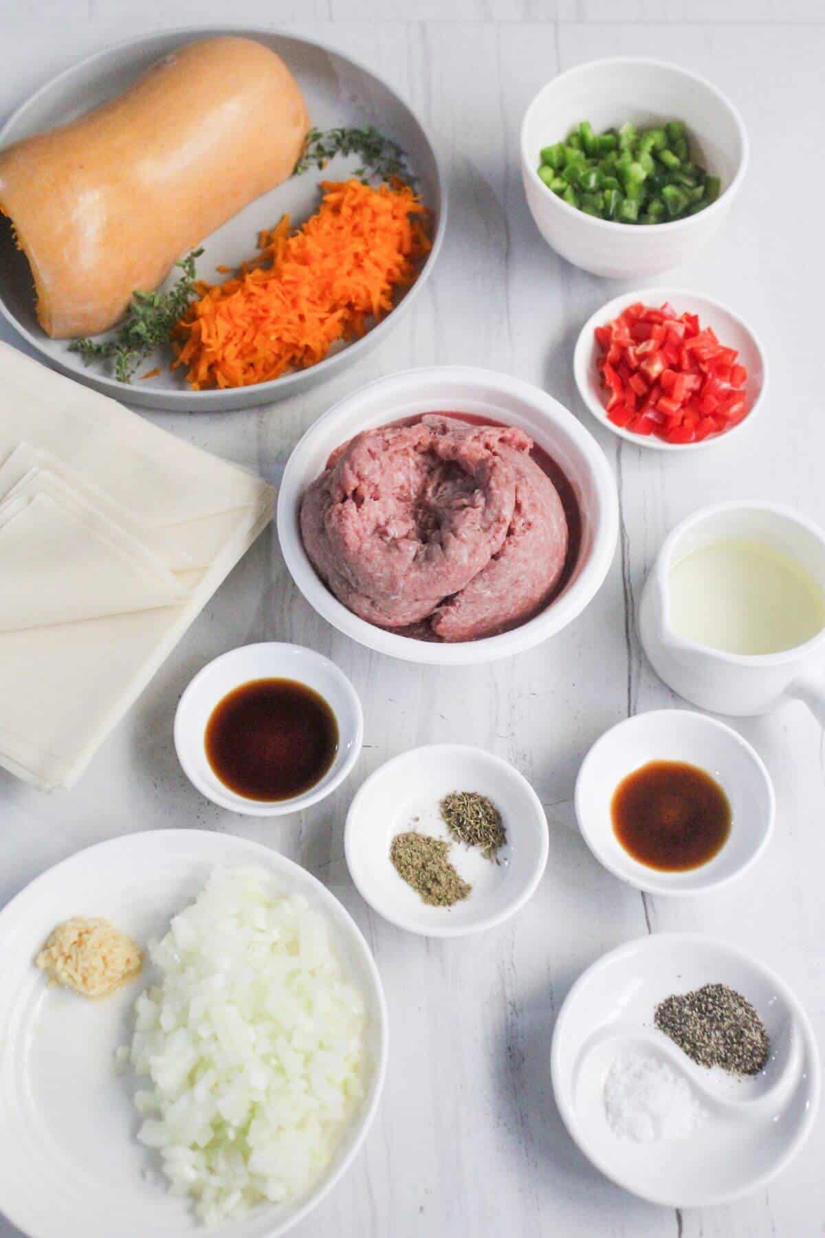 A flat lay of ingredients on a white surface includes ground meat, diced vegetables, seasonings, soy sauce, oil, and spring roll wrappers.