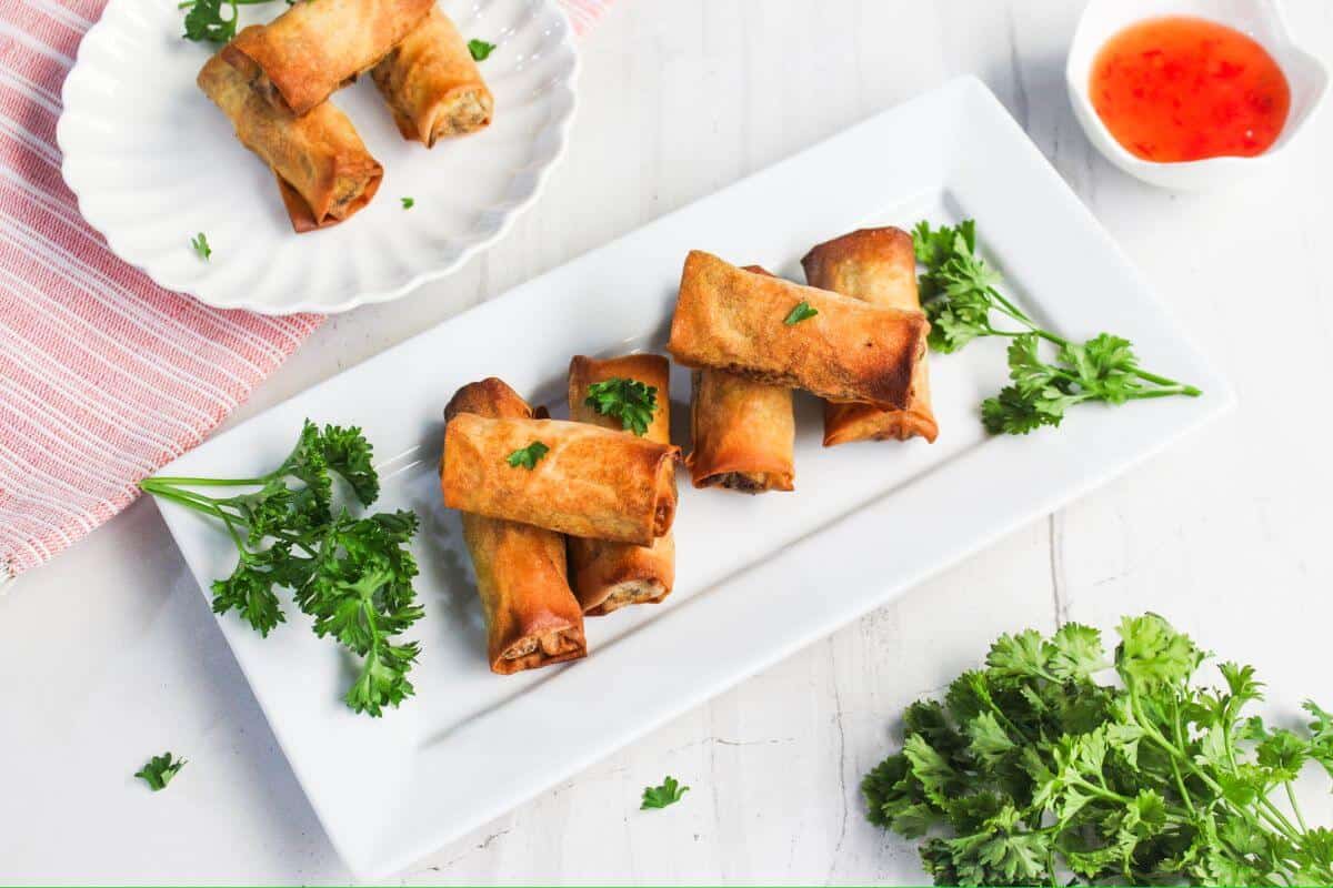 Crispy spring rolls on a rectangular plate with parsley garnish, accompanied by a small dish of dipping sauce.