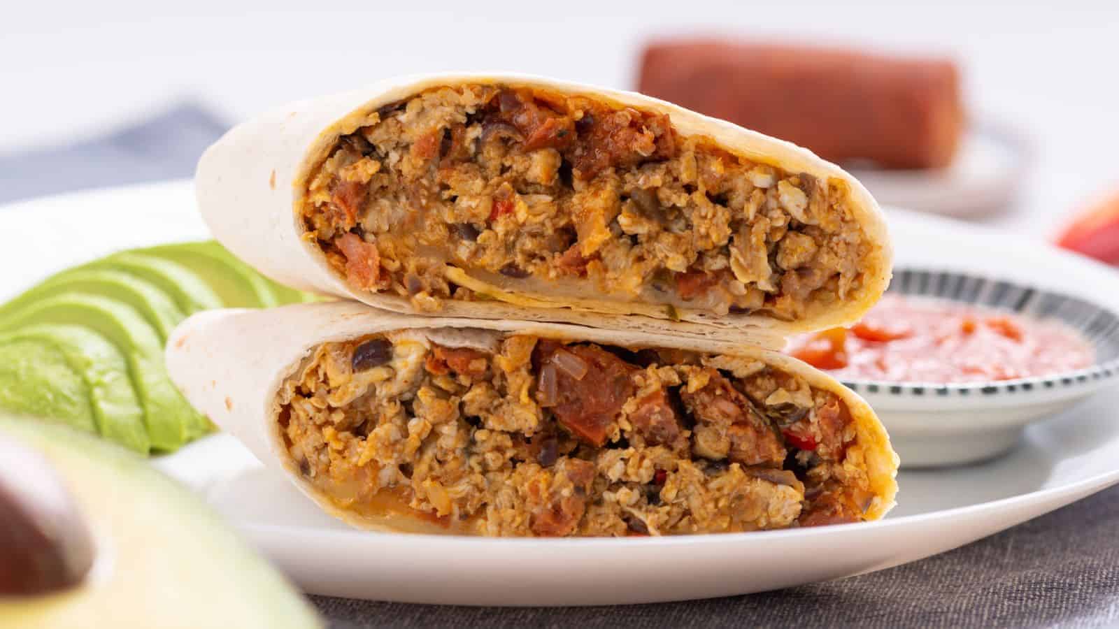 A close-up image of a sliced breakfast burrito filled with seasoned meat and eggs served on a white plate with avocado slices and dipping sauce in the background.